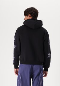 Sort hoodie med en stor hætte, prydet med lilla stjerne grafik på ærmerne. Blødt stof, afslappet pasform med ribstrikkede manchetter og kanten.