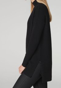 Svart stickad tröja med hög halsringning, ribbade ärmslut och sidofickor. Lång, lös design som sträcker sig till mitten av låret. Mjuk textur.