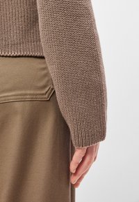 Gros plan sur le bras d'une personne portant un pull en maille marron et un pantalon marron texturé avec une couture visible sur la poche arrière.