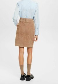 Vrouw in een beige suède rok, lichtblauw longsleeve shirt, tan sokken en zwarte instappers, staande met haar rug naar de camera.