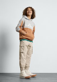 Felpa a blocchi di colore grigia e marrone con logo, pantaloni cargo beige con tasche laterali e sneakers alte bianche con suola marrone.