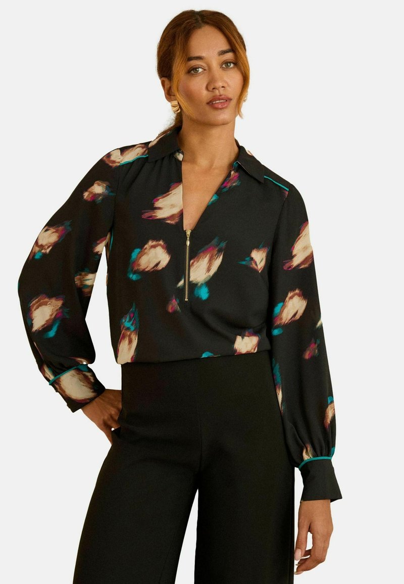 Fekete blúz absztrakt virágmintával bézs, türkiz és rózsaszín árnyalatokban; cipzáros gallérral, hosszú ujjakkal és laza szabással.