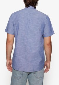 Camisa de manga corta, de color azul claro, hecha de una tela texturizada. Presenta un bajo recto y un pequeño cuello. Detalle de puntada en la parte posterior visible.