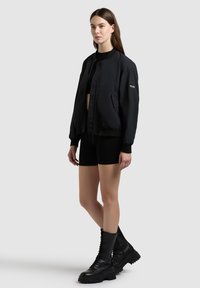 Femme debout portant une veste bomber noire, un crop top noir, un short ajusté noir et des bottes de combat à lacets noires sur un fond uni.