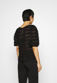 Svart genomskinlig topp med texturerade horisontella ränder, puffärmar och en nyckelhålsdetalj på baksidan. Kombinerad med svarta byxor.