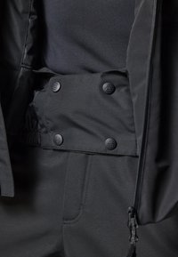 Chaqueta impermeable negra con cierre de botones a presión, cintura elástica y tejido liso con un sutil brillo. Incluye detalles en los bolsillos laterales.