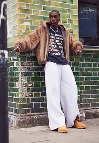 Överdimensionerad brun pälsjacka över en svart t-shirt med tryck, kombinerad med ljusgrå vida mjukisbyxor och tanfärgade stövlar mot en grön kakelvägg.