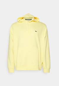 Felpa gialla con tasca a marsupio, cappuccio regolabile con lacci e un piccolo logo verde Lacoste sul petto. Tessuto morbido, design casual.