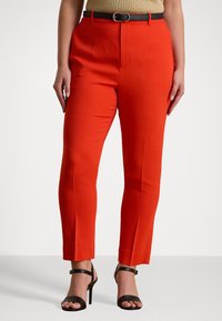 Pantaloni rossi su misura con pieghe frontali e taglio slim, abbinati a una cintura scura. Tacchi neri con cinturini completano il look.