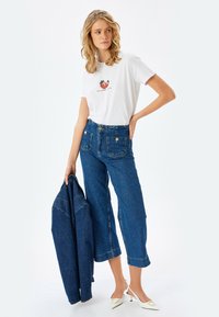 Hvid bomulds t-shirt med jordbær grafik, høj talje blå denim brede bukser, holder en mørk denimjakke, og hvide hæle.