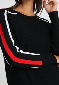 Pull en tricot noir à côtes, avec des manches longues ornées de bandes rouges et blanches le long des bras et un col rond.