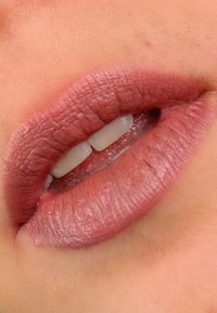 Matte mauve leppestift påført leppene, som viser en teksturert finish. Leppene er litt åpne og avslører hvite tenner.