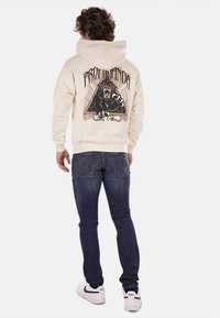 Felpa crema con una grafica di un leone nero sulla schiena, abbinata a jeans blu scuro e sneakers bianche. Presenta una tasca frontale e cappuccio con lacci.