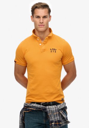 Orange polo shirt with a collared neck and three buttons, featuring a contrasting navy logo. Short sleeves with rolled cuffs, worn with a plaid shirt tied at the waist.