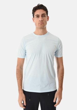 Giovane in piedi, indossa una maglietta a maniche corte azzurra e pantaloni neri, espressione neutra, sfondo bianco semplice.