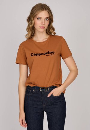 T-shirt en coton brun avec le texte "Cappuccino" en noir, associé à un jean bleu foncé et une ceinture noire. Coupe décontractée avec des manches courtes.