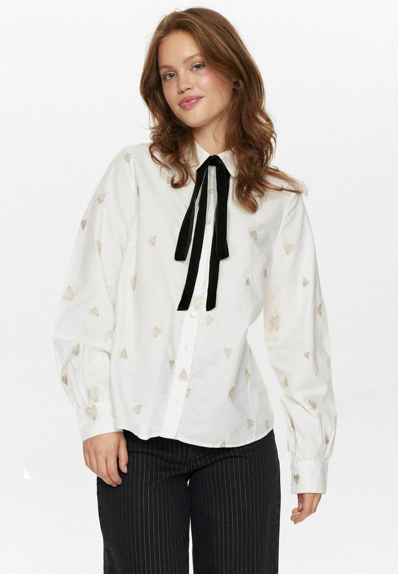 Witte blouse met kleine gouden hartpatronen, zwart strikje om de hals en lange mouwen met knoopsluitingen. Zwarte gestreepte broek.