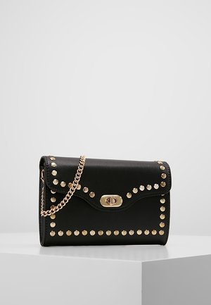 Zwarte leren clutch met gouden studs die de voorflap omlijnen, een gouden draaislotsluiting en een gouden kettingriem, gedrapeerd over een wit voetstuk.