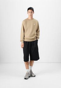 Beige Nike sweatshirt, zwarte shorts, grijze sneakers. De sweater heeft een ronde halslijn en ribgebreide mouwen, de shorts hebben een losse, casual pasvorm.