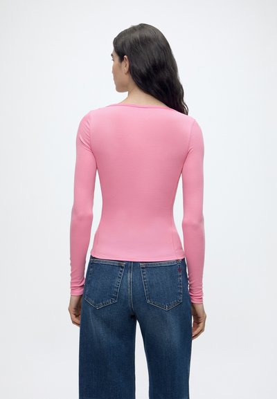 Femme debout de dos face à la caméra, portant un haut rose ajusté à manches longues et un jean bleu, sur un fond uni clair.