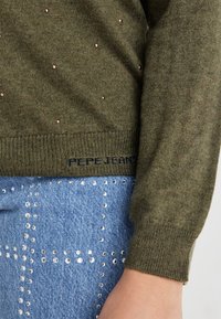 Maglione verde oliva con piccoli chiodi metallici, orlo a coste e logo "Pepe Jeans" sulla parte inferiore, abbinato a jeans blu con chiodi d'argento.