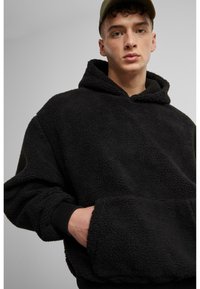 Sweat à capuche en polaire noire avec une surface texturée et douce, grande poche avant et poignets côtelés. Dispose d'une capuche avec cordon de serrage et d'une coupe décontractée.