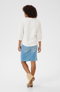 White top with eyelet balloon sleeves paired with a denim skirt. Model wears beige ankle boots, showcasing a relaxed, casual style.