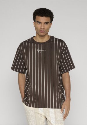 Muž s krátkymi kučeravými vlasmi, oblečený v hnedom tričku s vertikálnymi pruhmi a svetlých vzorovaných nohaviciach, stojí pred jednoduchým pozadím.