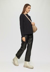 Schwarzes, strukturiertes Langarmoberteil, schwarze Lederhosen und beige Knöchelstiefel. Das Model trägt eine beige Umhängetasche und posiert in einer Gehhaltung.