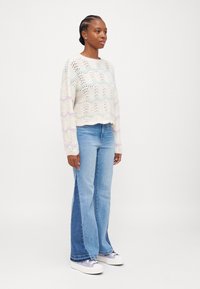 Pulôver de crochê em creme com padrões de ondas em tons pastéis, combinado com jeans azuis em estilo flare com um painel de denim mais escuro. Ténis brancos com pormenores em roxo.