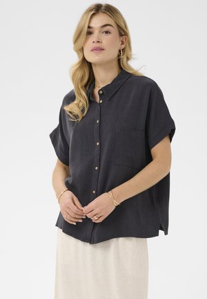 Szőke nő, laza sötétszürke rövid ujjú, gombos ingben és világos bézs szoknyában, összekulcsolt kézzel pózol.