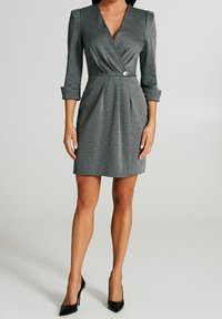 Szara, dopasowana sukienka w stylu kopertowym z rękawami o długości trzy czwarte, detalem guzikowym w talii oraz plisowaną fakturą, w komplecie z czarnymi szpilkami w szpic.