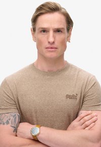 Camiseta de rayas marrón claro hecha de material suave, con mangas cortas y escote redondo. Tatuaje notable en el brazo izquierdo. Reloj dorado con esfera azul.