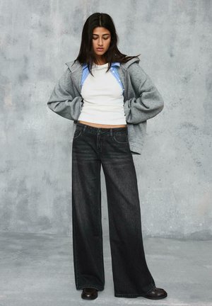 Jeune femme debout, mains sur les hanches, portant un jean noir à jambes larges, un débardeur blanc, une chemise rayée et un sweat à capuche gris, devant un mur gris.