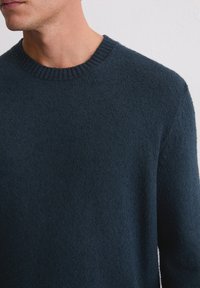 Donkerblauwe gebreide trui met een geribbelde ronde halslijn en zachte, gestructureerde stof; close-up van het bovenlichaam en het schoudergebied.