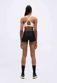 Γυναίκα που  στέκεται και κοιτάζει προς τα πίσω, φορώντας λευκό αθλητικό σουτιέν με σταυρωτή πλάτη, μαύρα σορτς Under Armour, μαύρες κάλτσες και μαύρα αθλητικά παπούτσια Under Armour.