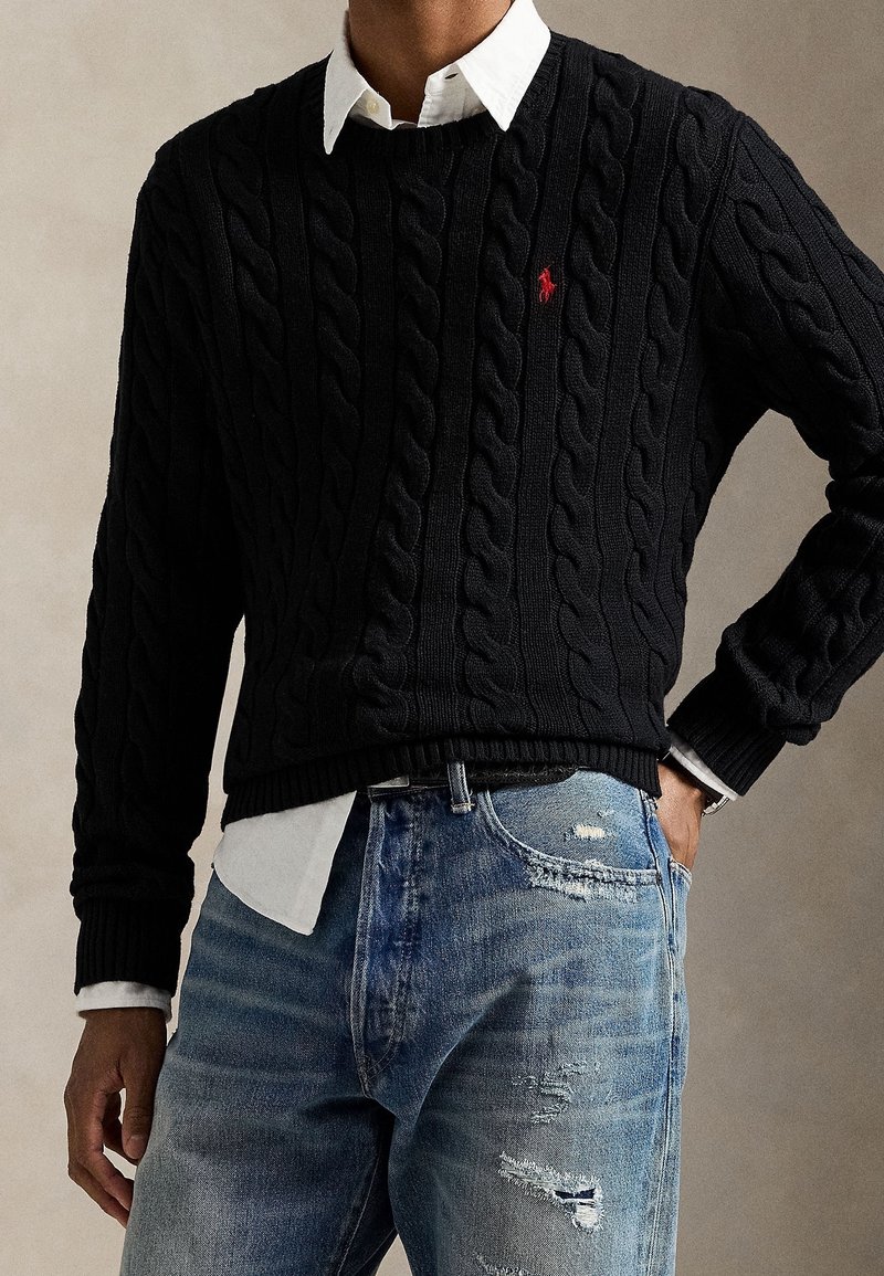 Homme portant un pull en maille torsadée noire par-dessus une chemise blanche, associé à un jean bleu déchiré, main dans la poche sur un fond neutre.