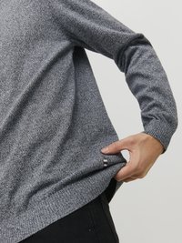 Grijze gebreide trui met lange mouwen, een relaxte pasvorm en een klein decoratief label nabij de zoom. Zichtbare hand die de stof aanpast.