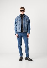 Džinsinis švarkelis su priekinėmis kišenėmis ir juodu užtrauktuku, dėvimas ant juodo golfo, derinamas su mėlynais džinsais ir juodais batais.