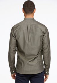 Camisa de manga larga en verde oliva oscuro, hecha de tela texturizada con cuello abotonado y mangas con puños, que presenta un pliegue central en la parte posterior.