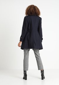Cappotto di lana blu con una forma aderente, maniche con bottoni e orlo svasato. Abbinato a pantaloni a quadri e stivali neri con tacco. 