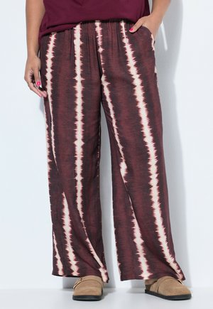 Persona che indossa pantaloni larghi a righe marroni e crema, scarpe slip-on marroni e una maglia color mogano, in piedi contro una parete chiara.