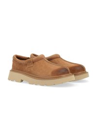 Scarpe slip-on in suede marrone con suola in gomma testurizzata, caratterizzate da una punta arrotondata e dettagli di cucitura discreti. Etichetta con logo sul lato.