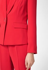 Czerwony marynarski żakiet o dopasowanym kroju z klapami, pojedynczym srebrnym guzikiem i dwiema przednimi kieszeniami. Materiał wygląda na gładki i sztywny.
