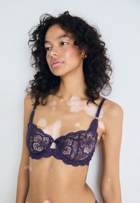 Soutien-gorge en dentelle violette avec motif floral, design semi-transparent, bretelles ajustables et un petit nœud central. Texture lisse et bords festonnés.