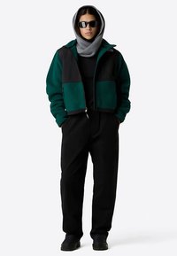 Zelená a černá fleecová bunda na zip, volnějšího střihu, nošená přes černý top. Černé kalhoty a šedý šátek, doplněné tmavými slunečními brýlemi.
