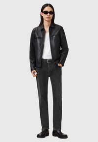 Chaqueta de cuero negra con cuello, camiseta blanca, pantalones de mezclilla gris, cinturón negro y botas de cuero negras. Modelo lleva gafas de sol oscuras.