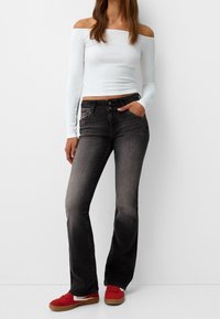 Bílý top s odhalenými rameny a dlouhými rukávy v kombinaci se šedými rozšiřujícími se džíny. Džíny mají přední kapsy a lehce vybledlý vzhled. Červené boty doplňují celý outfit.