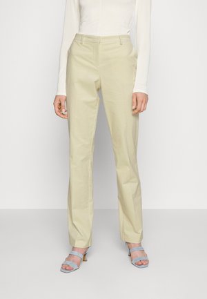 Persoon draagt beige rechte broek, wit shirt met lange mouwen en lichtblauwe open-toe hakken tegen een effen achtergrond.