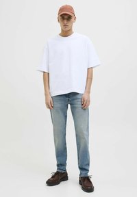 T-shirt blanche oversized, jean bleu clair, chaussures à lacets marron, et une casquette couleur rouille avec du texte en relief. Le mannequin pose devant un fond uni.